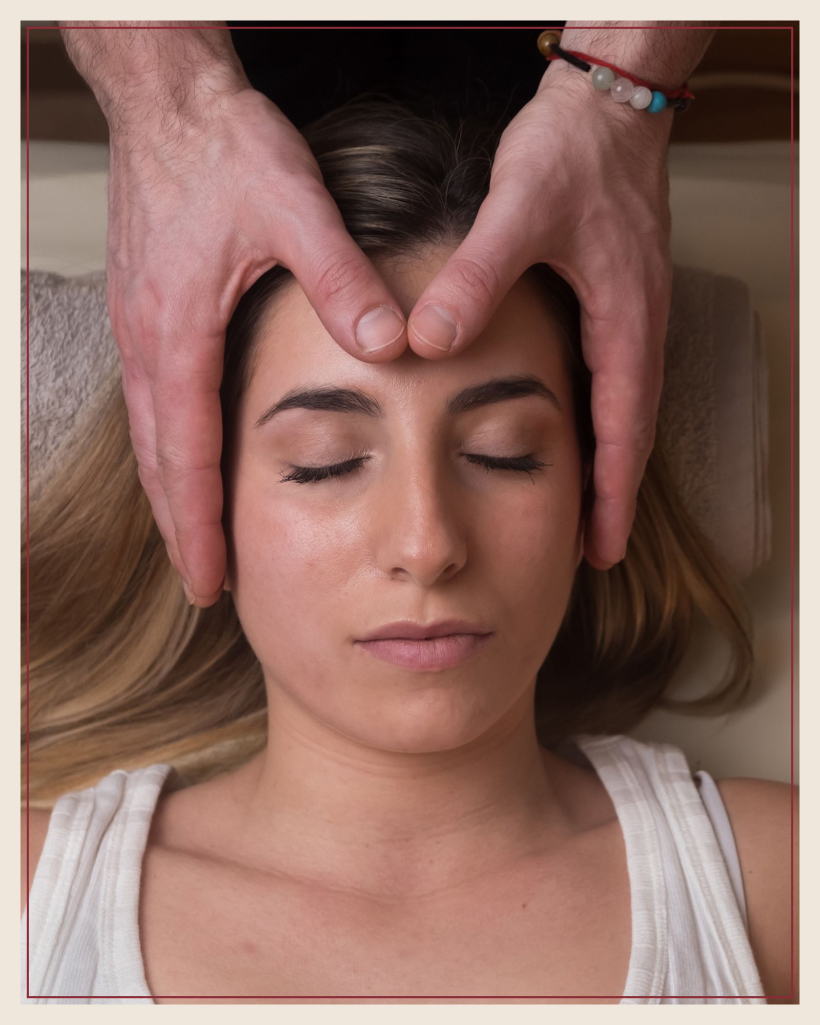 A practitioner's hands resting lightly at a client's forehead as she lies face up, eyes closed, wearing a white top