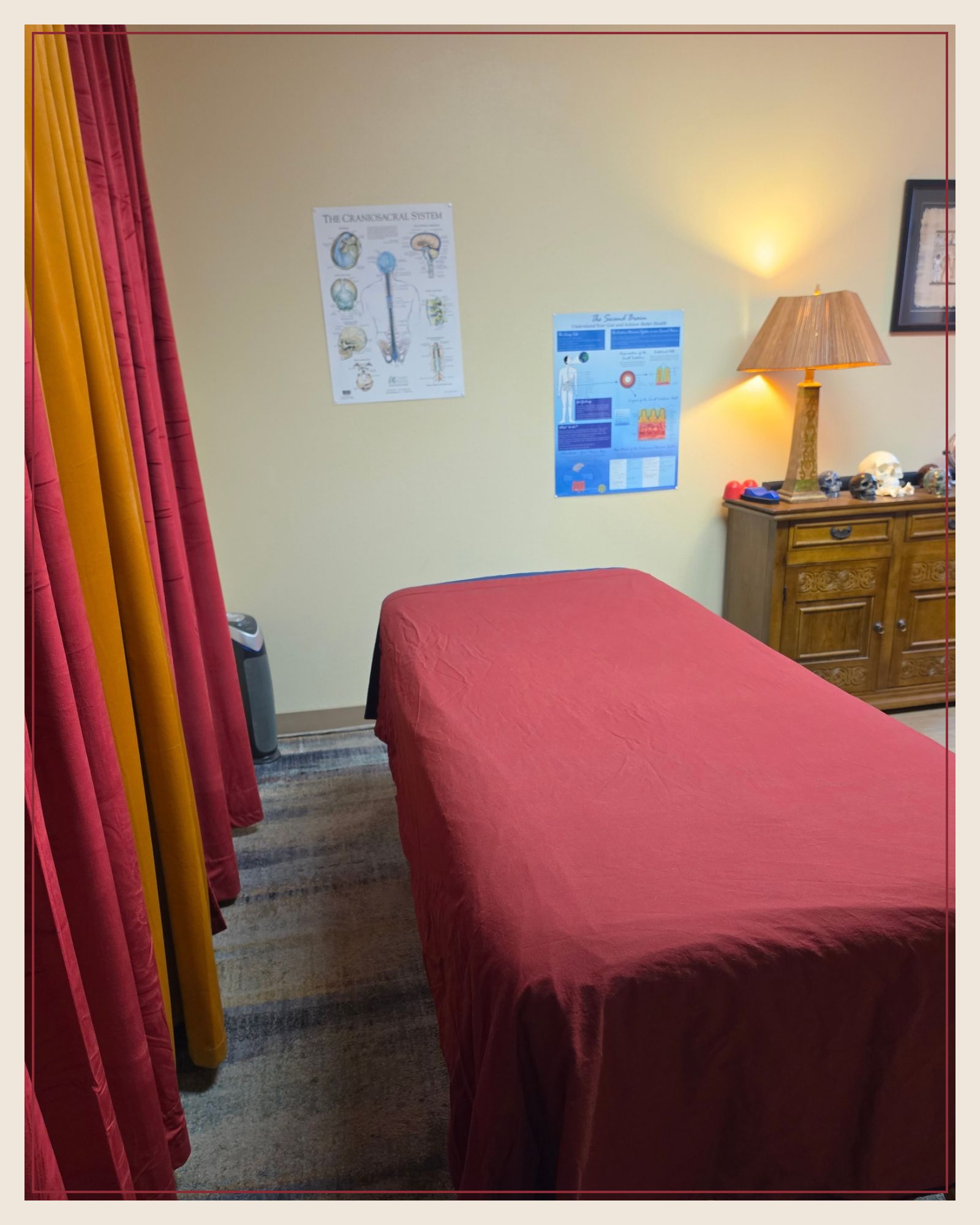 The treatment room: a massage table with a red sheet, anatomy posters on the wall, and a warm lamp on a wooden cabinet.